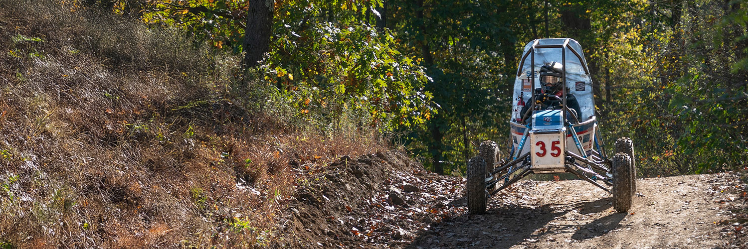 A dirt path winds through a forest with dense green and yellow foliage. Sunlight filters through the trees, casting dappled shadows on the ground. The edge of a vehicle tire is visible on the right side of the image.